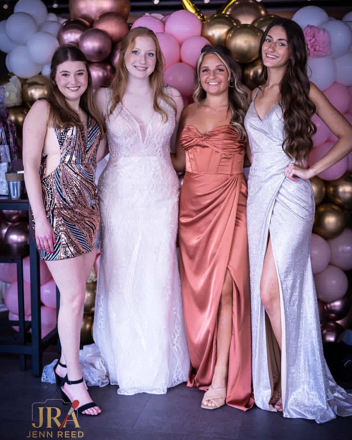 Four Young Women Pose Together In Elegant Dresses Against A Backdrop Of Colorful Balloons, Celebrating A Special Occasion.