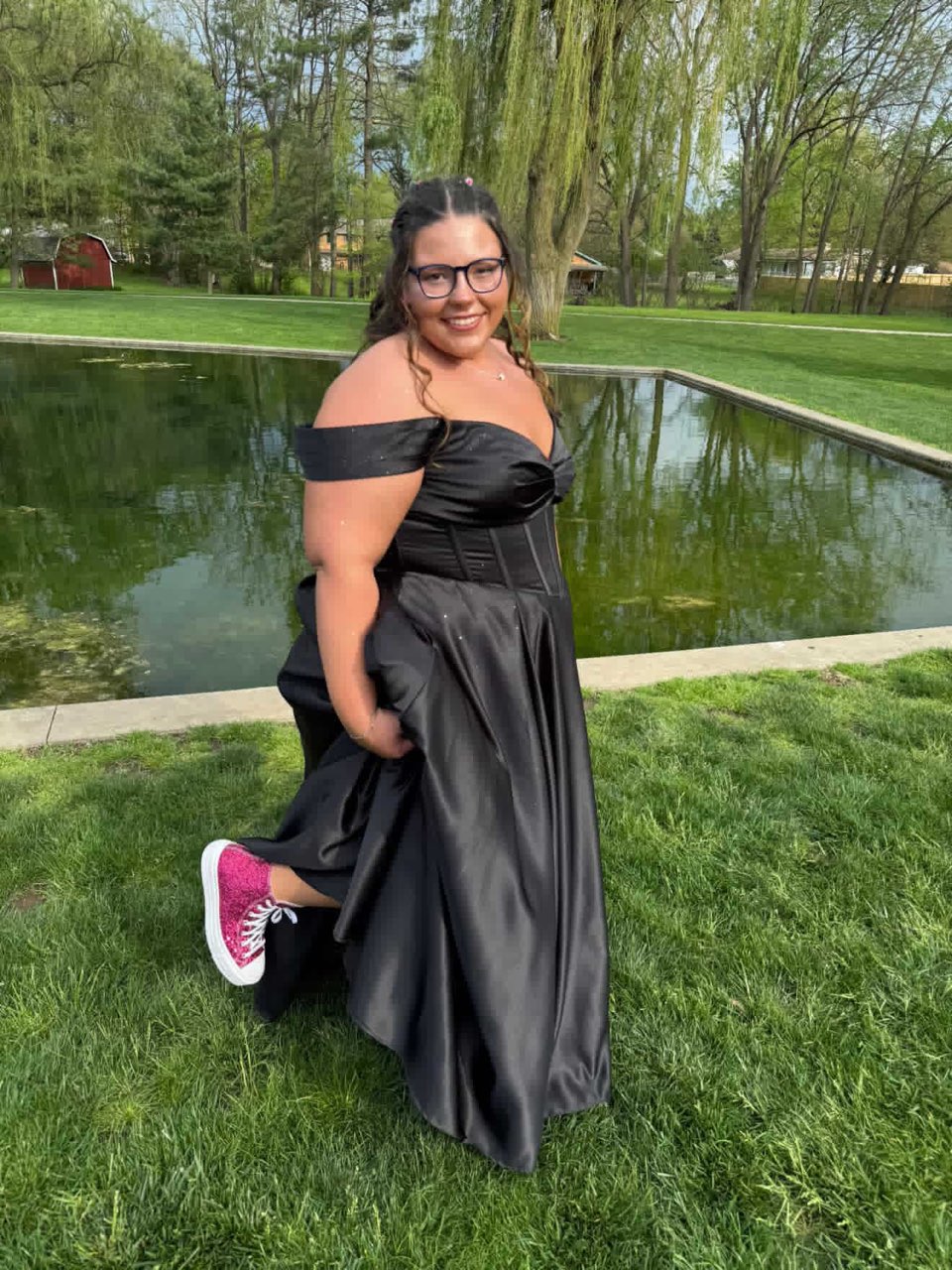 A Young Woman In A Sleek Black Dress Playfully Poses By A Pond, Wearing Vibrant Pink Sneakers. Lush Greenery And Trees Provide A Scenic Backdrop.