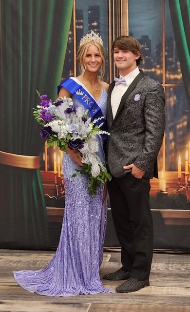 A Crowned Queen In A Sparkling Lavender Gown Holds A Bouquet, Standing Beside A Formally Dressed Young Man, Both Smiling Against A Romantic Backdrop.