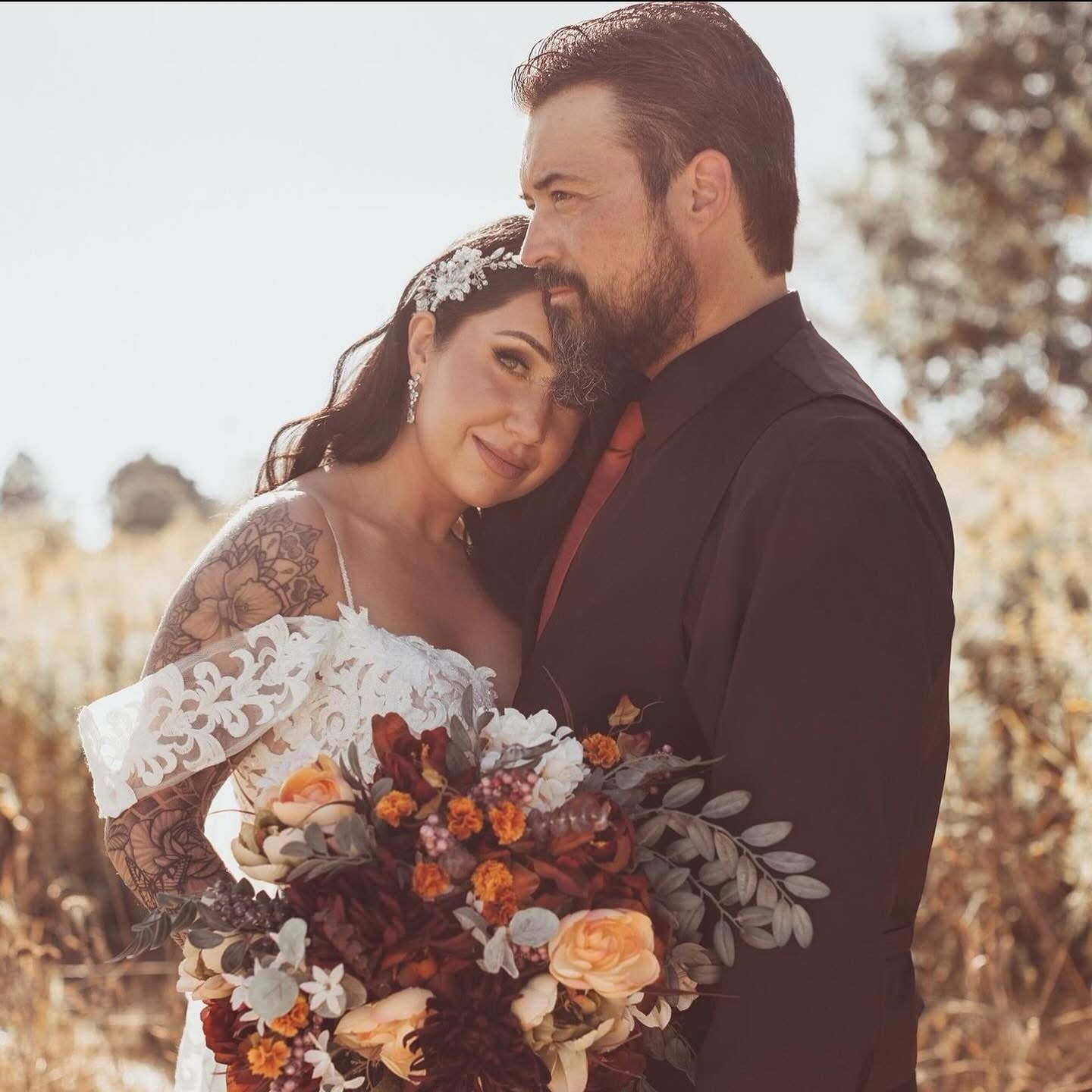 A Bride With Intricate Tattoos Wears A Lace Gown, Holding A Colorful Bouquet, As She Embraces Her Partner In A Sunlit Field, Capturing A Romantic Moment.