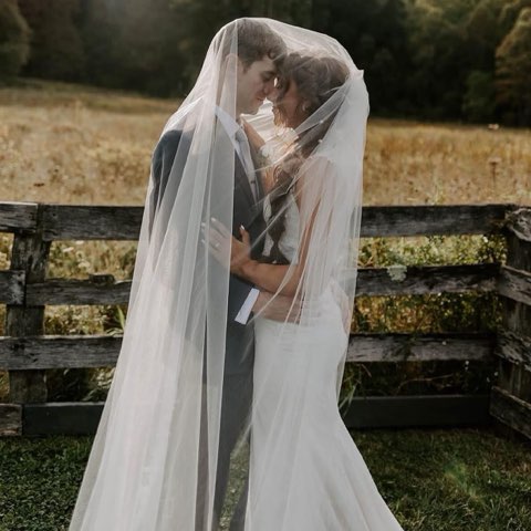 A Bride And Groom Share A Romantic Moment Under A Veil In A Rustic Outdoor Setting, Surrounded By A Grassy Field And Wooden Fence.