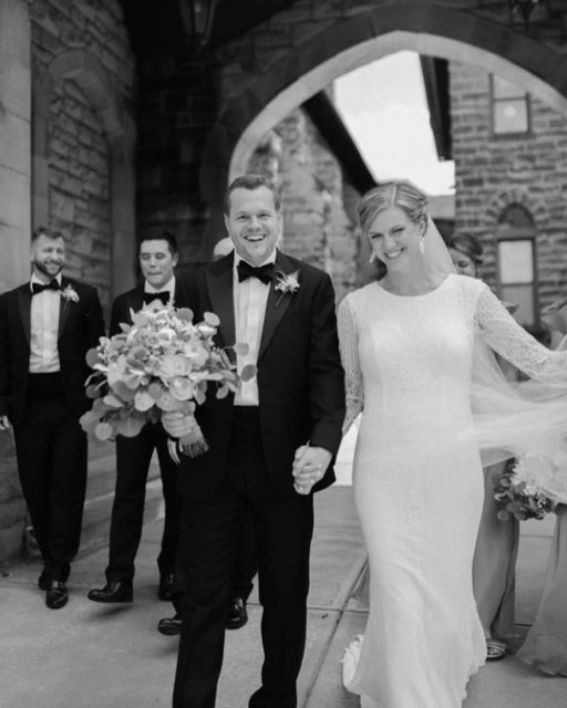 Joyful Bride And Groom Walk Hand In Hand, Surrounded By Wedding Party In Elegant Attire, Under A Beautiful Stone Archway.