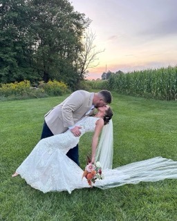 Newlyweds Share A Romantic Dip Kiss In A Lush Green Field At Sunset, Surrounded By Cornfields And A Colorful Bouquet.