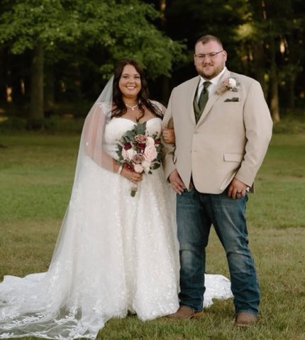 Bride In A Lace Wedding Gown Holds A Bouquet, Standing Beside The Groom In A Beige Suit And Jeans. They Smile In A Lush Outdoor Setting.