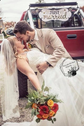 A Bride In A White Gown Leans Back, Embraced By Her Groom In A Tan Suit, Both Smiling Joyfully. A "Just Married" Sign Hangs From A Red Truck Behind Them, Capturing Their Celebratory Moment.