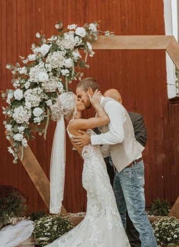 A Bride In A Lace Gown And Groom In A Vest Kiss Under Floral Decor At Their Wedding Ceremony, Set Against A Rustic Red Barn Backdrop.