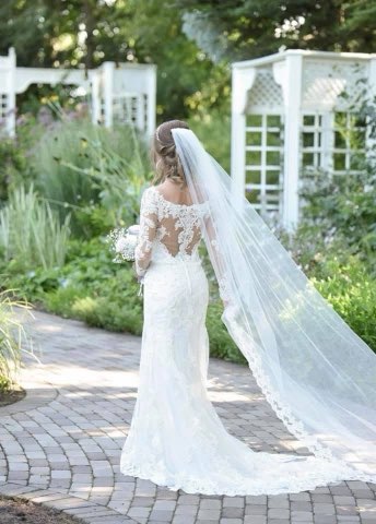 A Bride In A Lace Gown And Flowing Veil Walks Down A Garden Path, Surrounded By Lush Greenery And Floral Accents, Embodying Wedding Elegance.