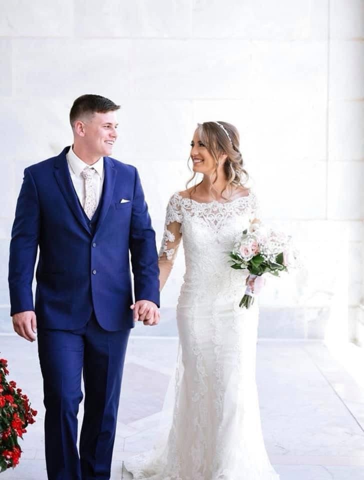 Smiling Bride In A Lace Gown Holds A Bouquet As She Walks Hand-in-hand With The Groom In A Navy Suit, In A Bright Outdoor Setting.