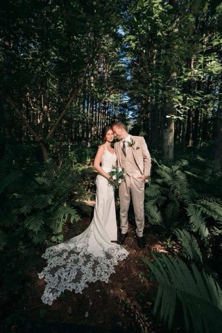 Bride And Groom Pose Lovingly In A Lush Forest, Surrounded By Greenery. The Bride Wears A Lace Gown While The Groom Is Dressed In A Beige Suit.