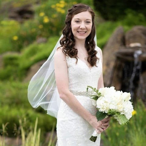Bride In A Lace Wedding Dress Holding A Bouquet Of White Flowers, Smiling In A Lush Outdoor Setting With Greenery And A Water Feature In The Background.