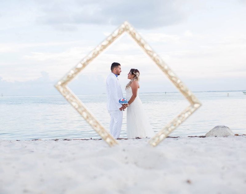 Couple In Wedding Attire Stand Holding Hands On A Beach, Framed By An Ornate Golden Picture Frame, With A Serene Ocean Backdrop.