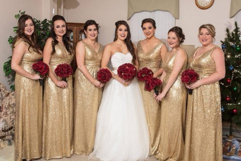 Bridesmaids In Gold Sequin Dresses Stand Smiling With The Bride In A White Gown, All Holding Red Bouquets. A Festive Decor Enhances The Joyful Atmosphere.