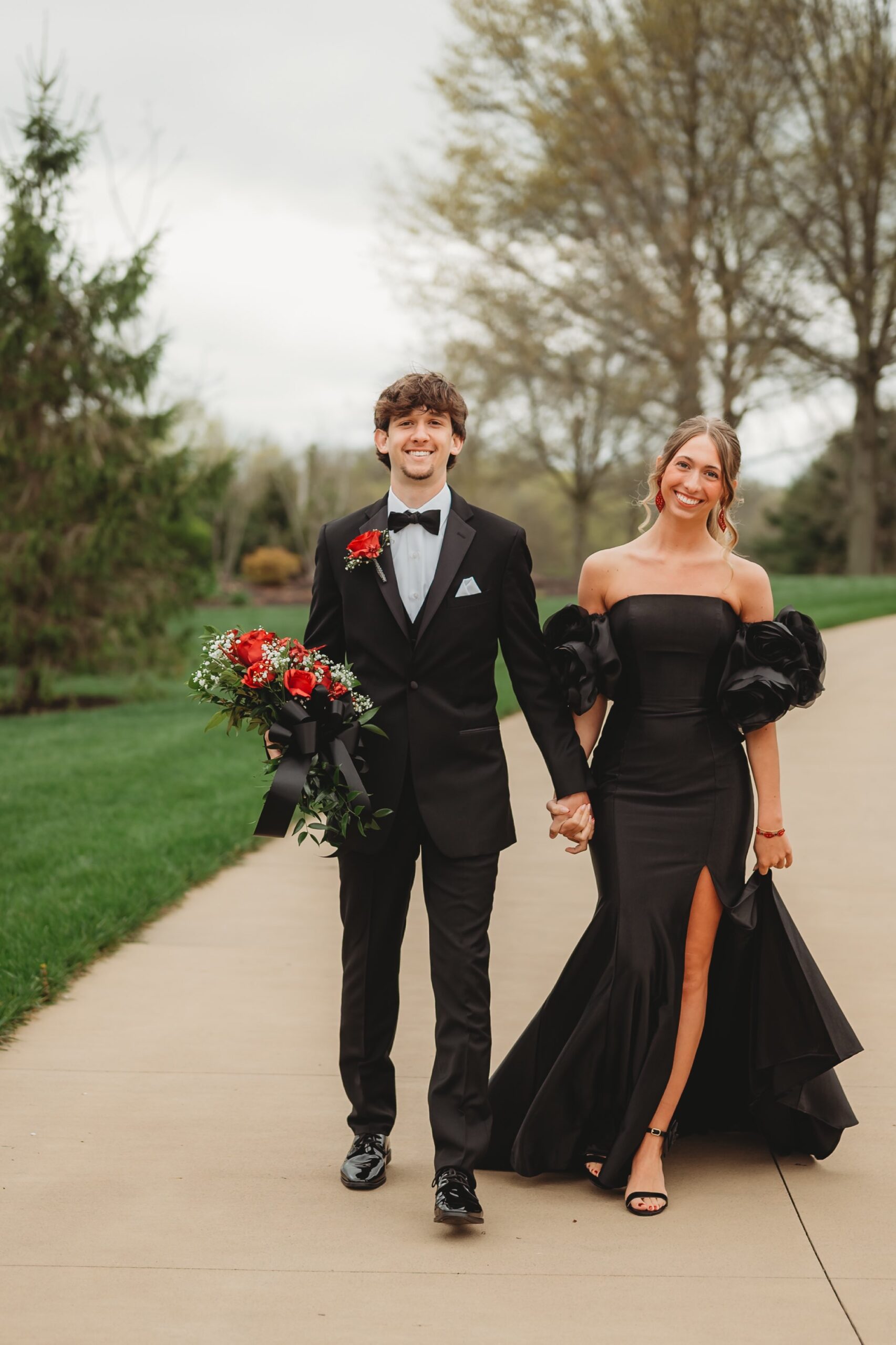 A Young Couple Walks Hand-in-hand Along A Paved Path, Dressed Elegantly For A Formal Event. The Woman Wears A Striking Black Gown, And The Man Sports A Classic Tuxedo With A Rose Boutonniere.