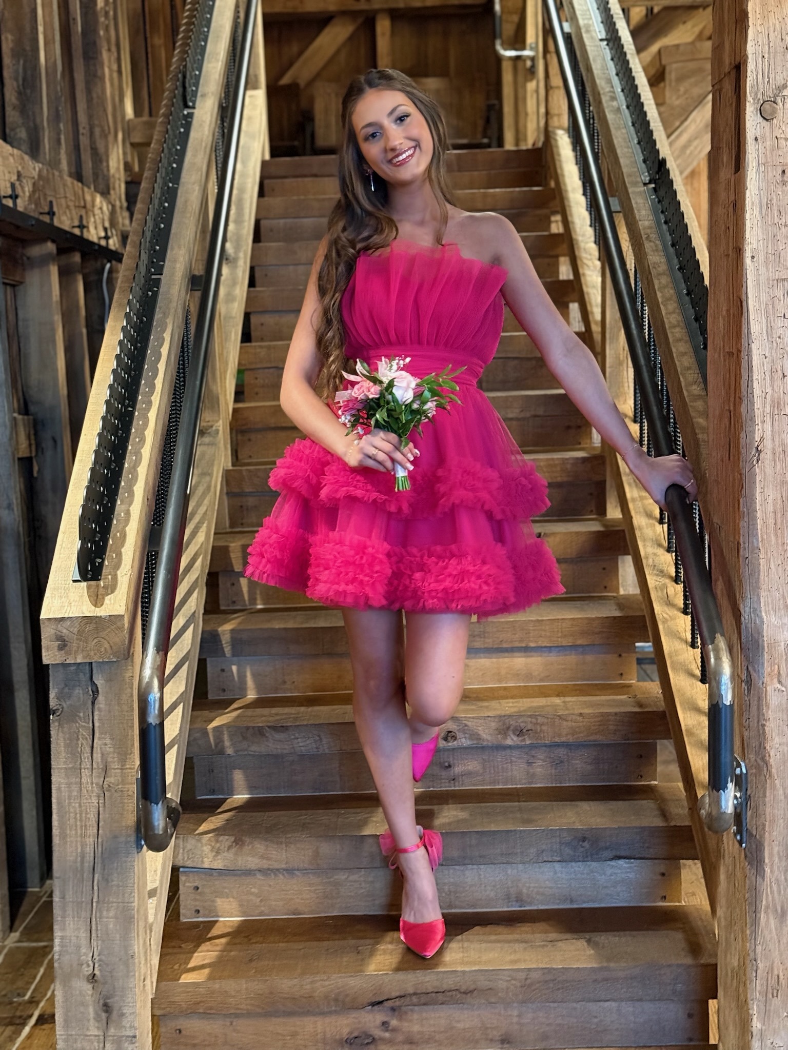 A Young Woman Wearing A Vibrant Pink Dress Poses On A Wooden Staircase, Holding A Bouquet Of Flowers, Smiling Confidently.