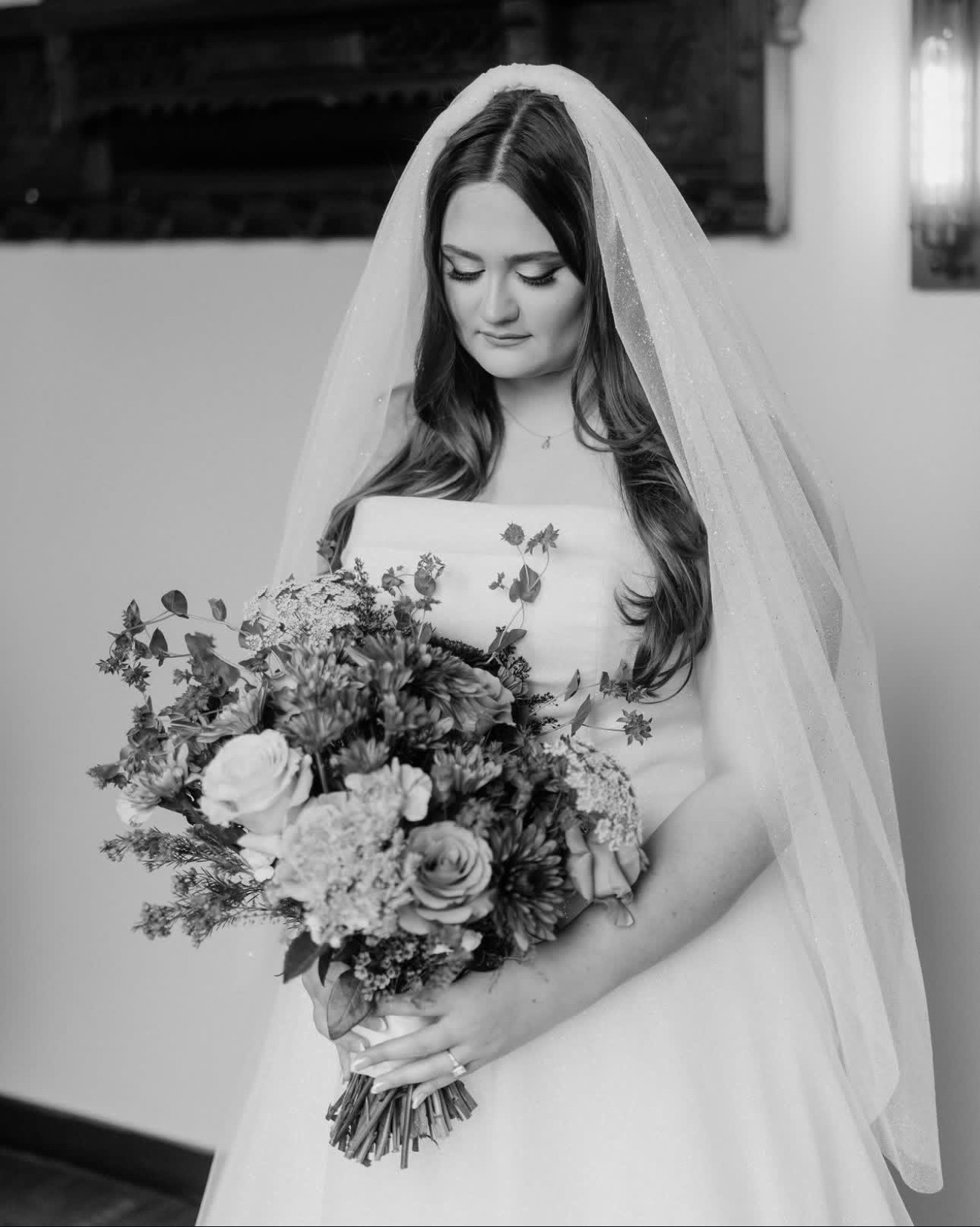 Bride In A White Gown And Veil Gazes Down At Her Floral Bouquet, Showcasing A Moment Of Reflection Before The Wedding Ceremony.