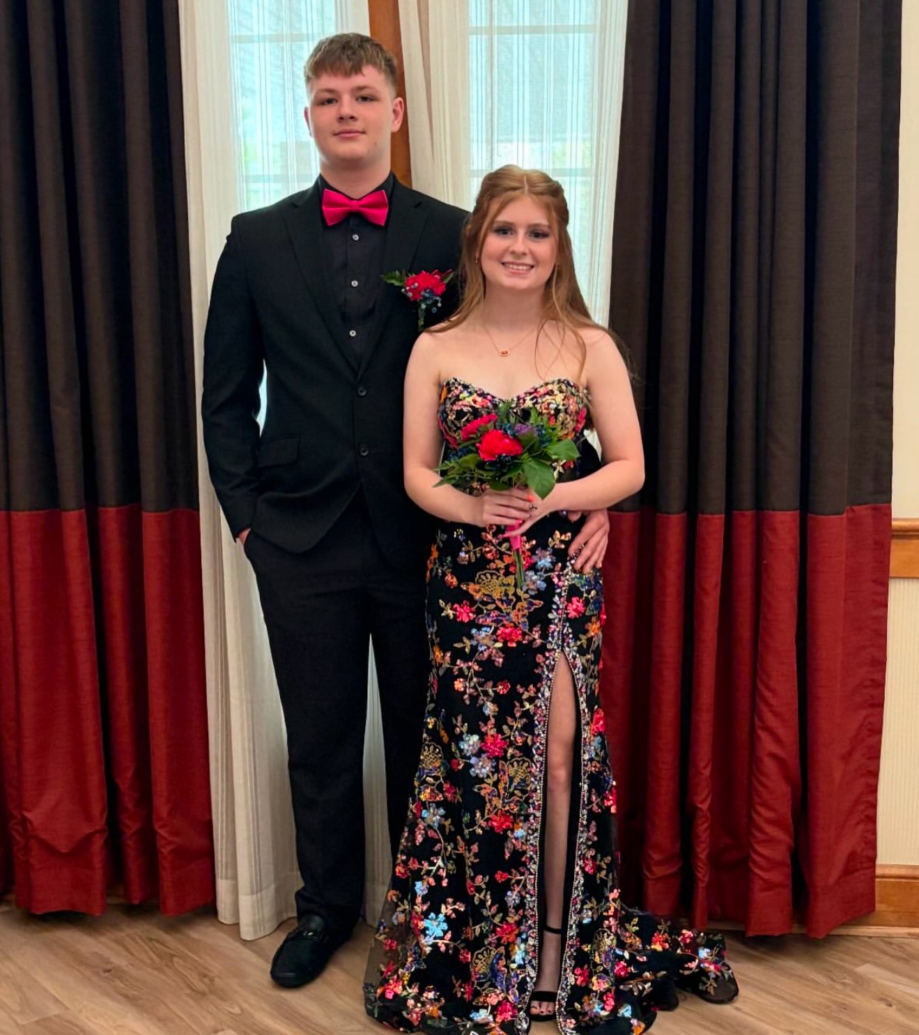 A Couple Poses For A Formal Photo, The Girl In A Floral Black Dress Holding A Bouquet, The Boy In A Black Suit With A Pink Bow Tie.