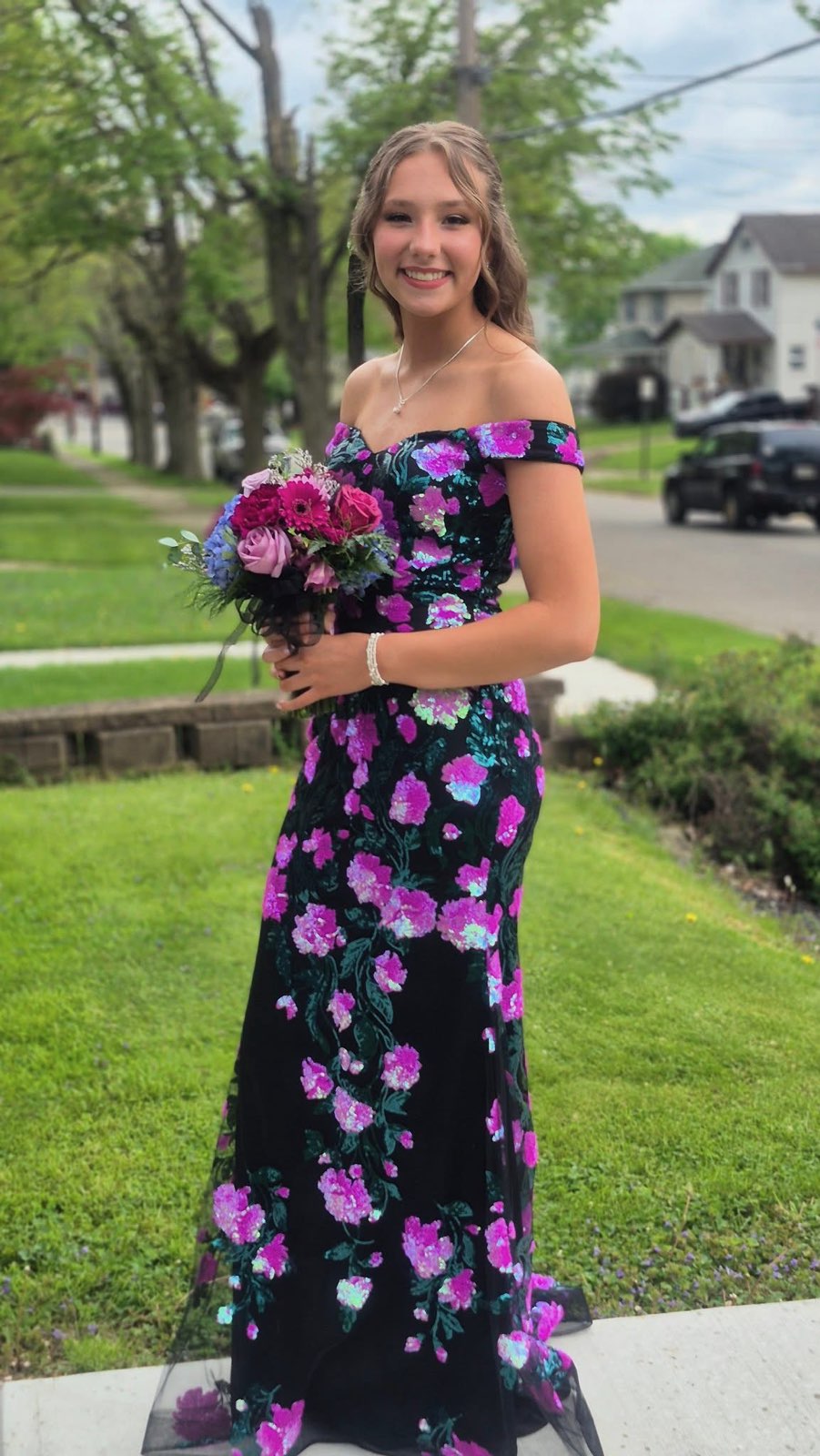 A Young Woman In A Floral Off-the-shoulder Dress Smiles While Holding A Bouquet, Standing In A Green Yard On A Sunny Day.
