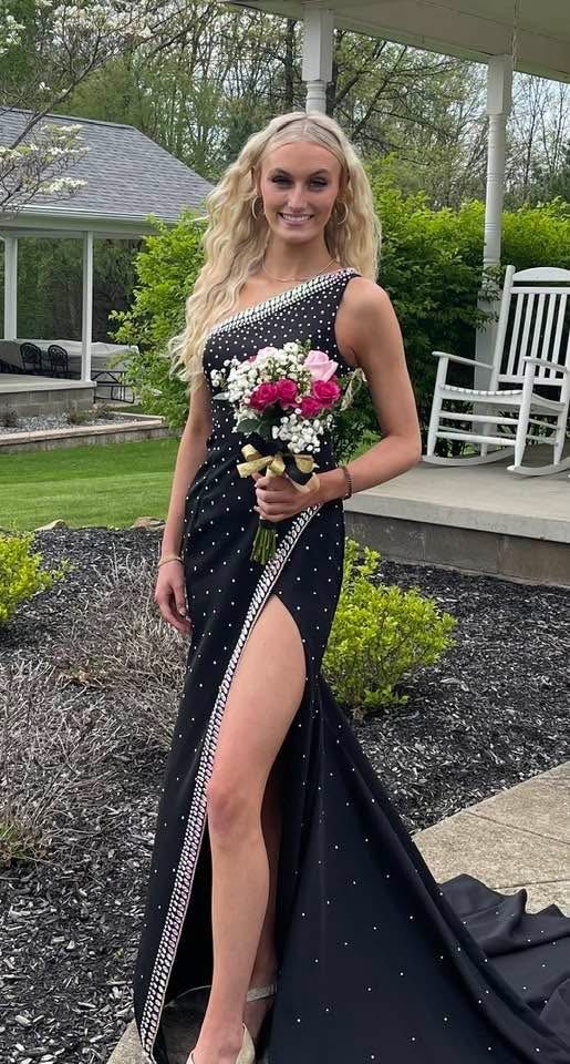 A Young Woman Poses In A Stunning Black Gown Adorned With Rhinestones, Holding A Bouquet Of Pink And White Flowers, Against A Garden Backdrop.