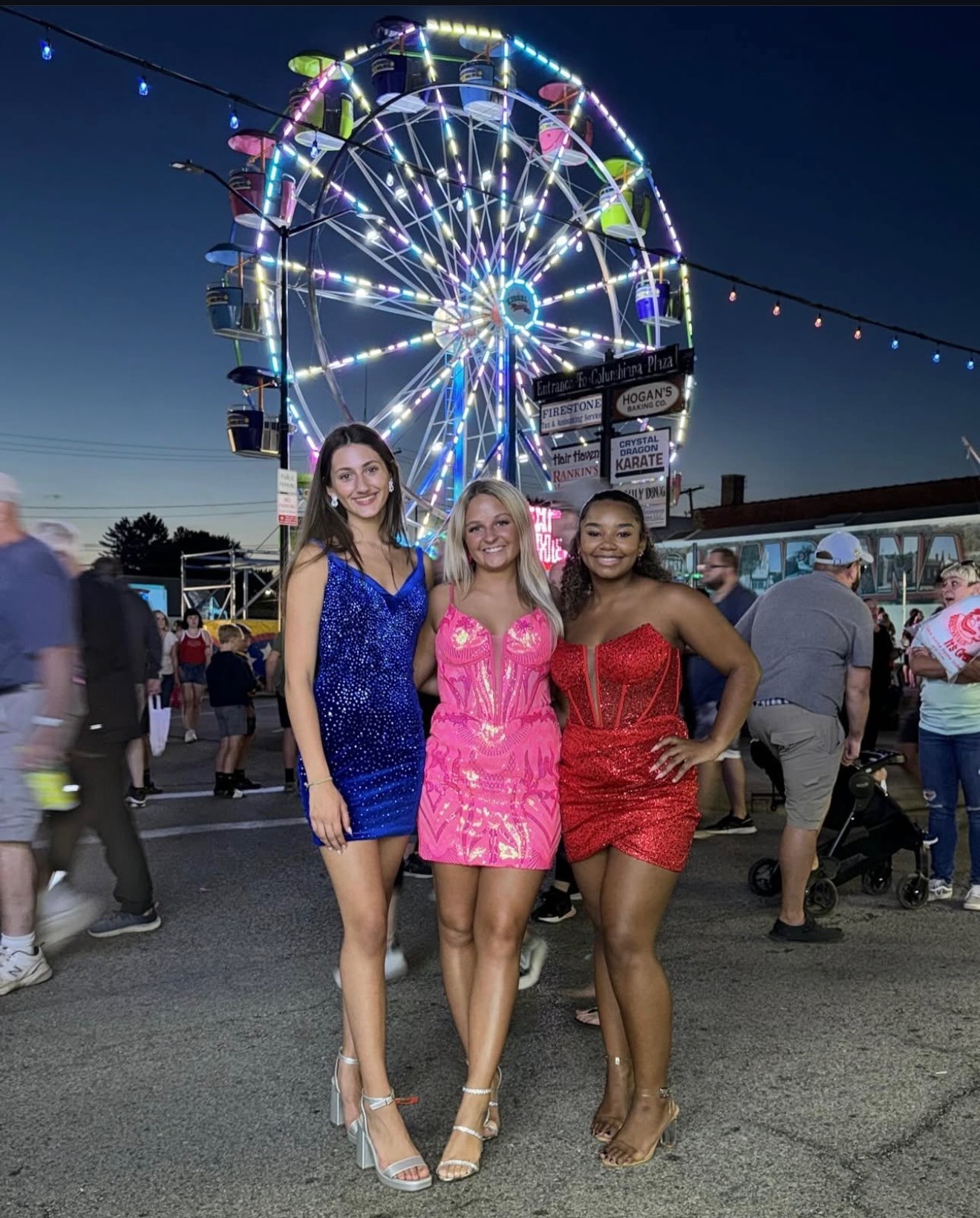 Three Young Women In Vibrant, Sparkly Dresses Pose Together In Front Of A Brightly Lit Ferris Wheel At A Fair, Capturing A Fun Summer Evening.