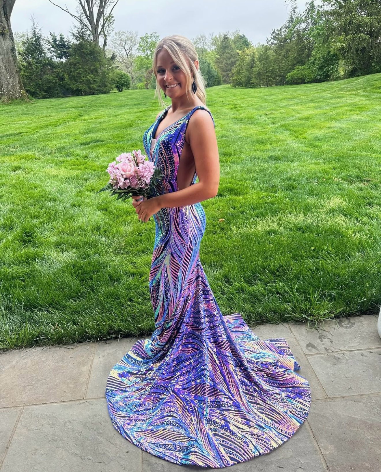 A Woman Stands In A Colorful, Shimmering Gown With A Train, Holding A Bouquet Of Pink Flowers. She Smiles Against A Lush Green Backdrop.