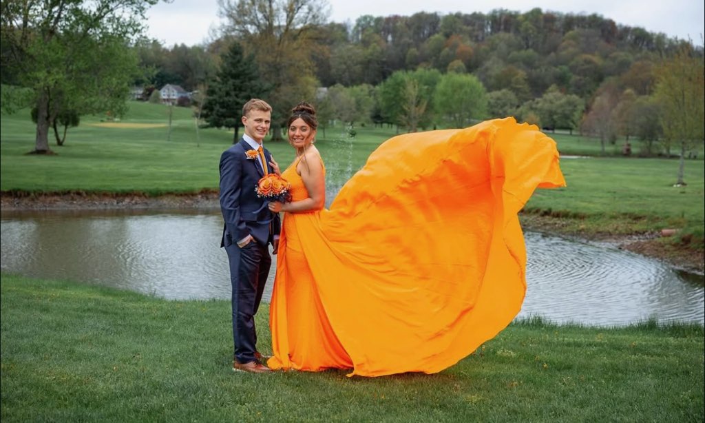 A Couple Poses By A Serene Pond, The Woman In A Flowing Orange Gown, Symbolizing Celebration And Romance, Surrounded By Lush Greenery.