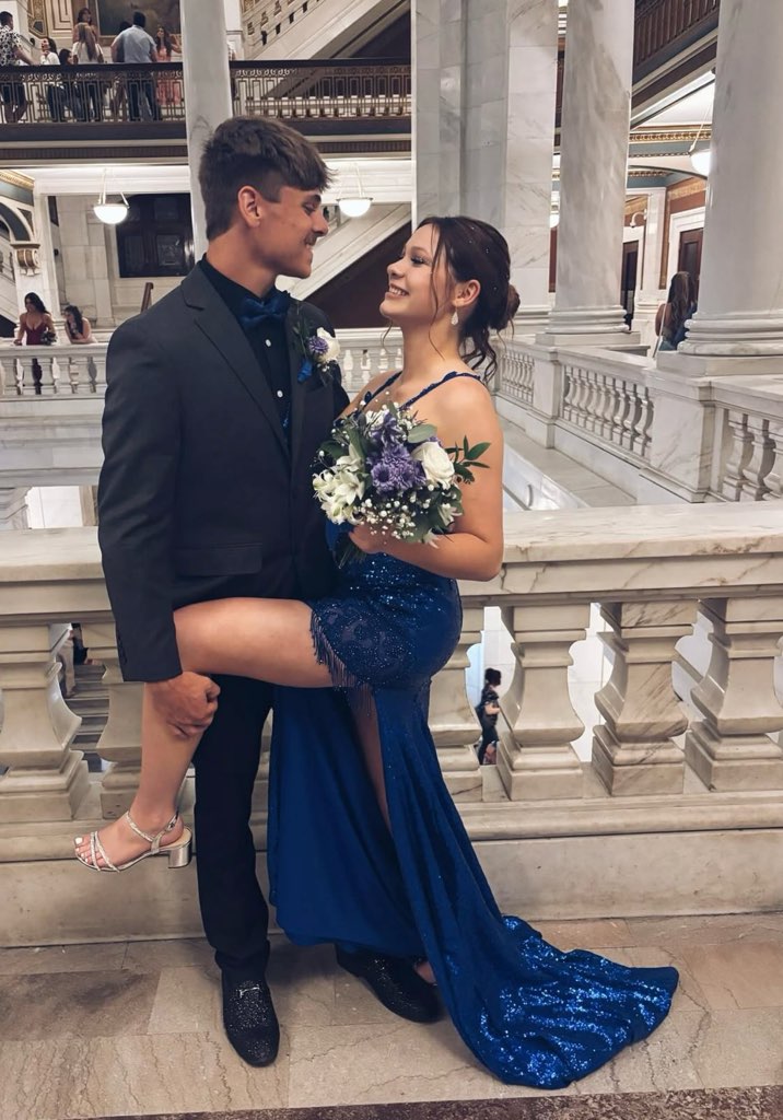 A Young Couple Smiles At Each Other In A Grand, Marble Hall, Dressed For A Formal Event; She Wears A Sparkling Blue Gown And Holds A Bouquet.