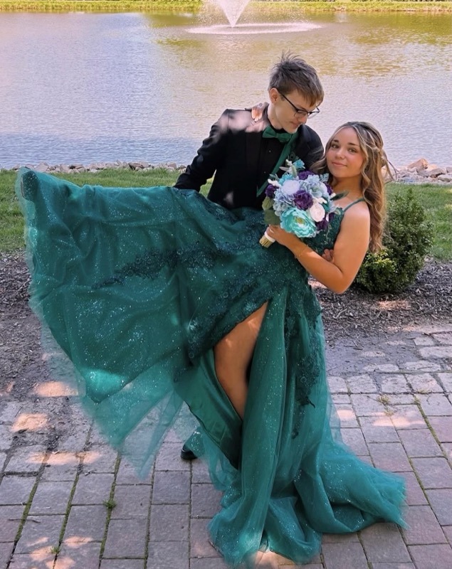 A Young Couple Poses By A Pond; The Girl In A Sparkling Teal Gown Holds A Bouquet, While The Boy, In A Tuxedo, Lifts Her Dress Playfully.