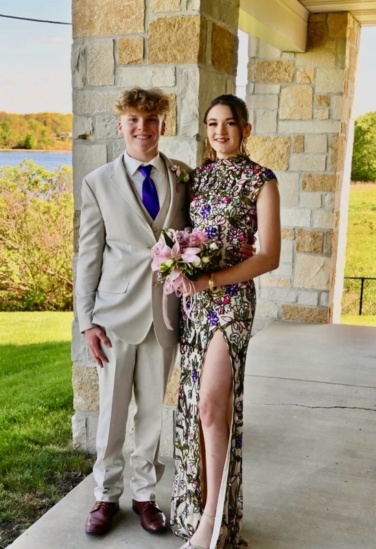 A Couple Poses For A Photo Before An Event, The Young Man In A Light Suit And Purple Tie, And The Woman In A Floral Dress Holding A Bouquet.